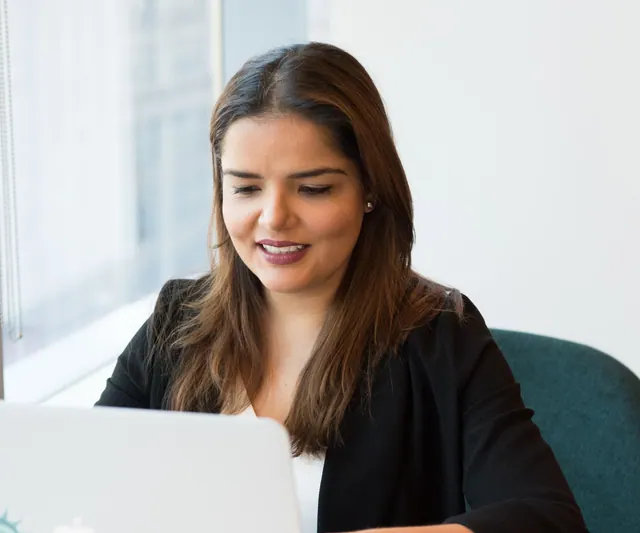 Woman working on a laptop beside a bright office window.